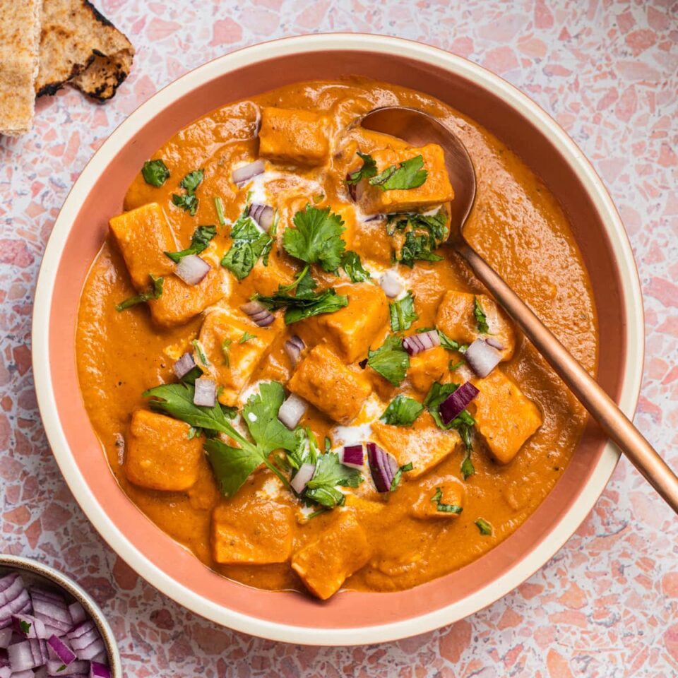 Bowl of butter paneer topped with cilantro and red onions with naan in the background.