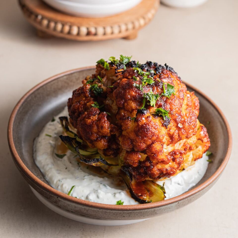 A whole roasted cauliflower with harissa glaze set atop yogurt in a bowl.