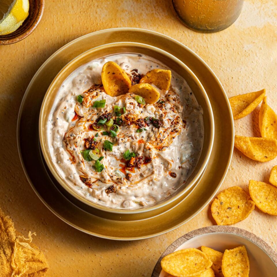 A bowl of sour cream dip with a bowl of chips next to it and chips spilled on the counter.