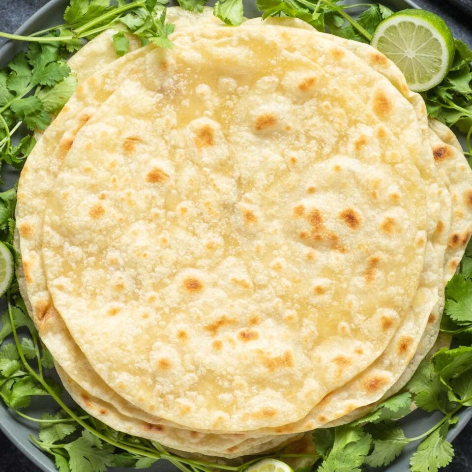 Vegan tortillas stacked up on a plate with fresh cilantro and lime.