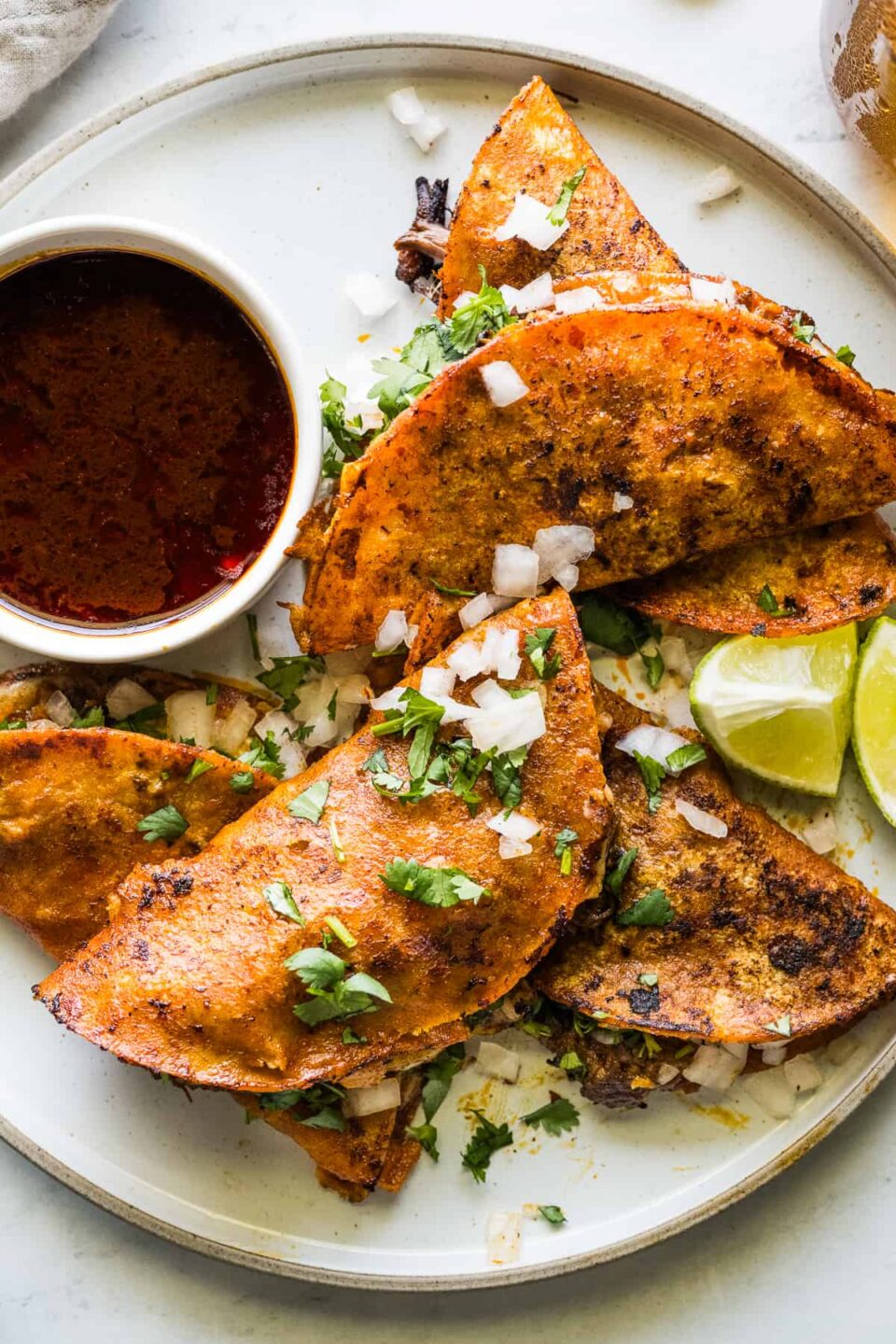 Birria tacos on a plate served with birria consomé and topped with diced white onion, cilantro, and limes.