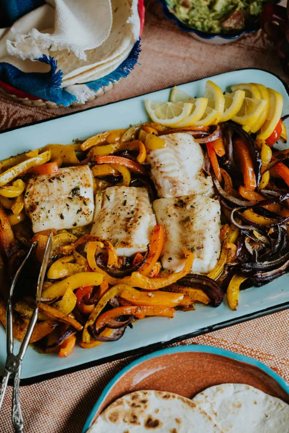 flat lay shot of a rectangular pale blue serving platter with cod and fajita veggies next to a plate of warm flour tortillas for making sheet pan fish fajitas.