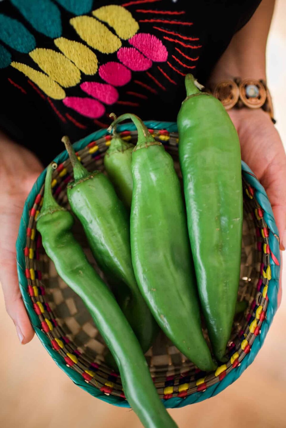 woman in a black shirt embroidered with bright pink, yellow, and turquoise holding a colorful woven rattan bowl with fresh anaheim peppers.