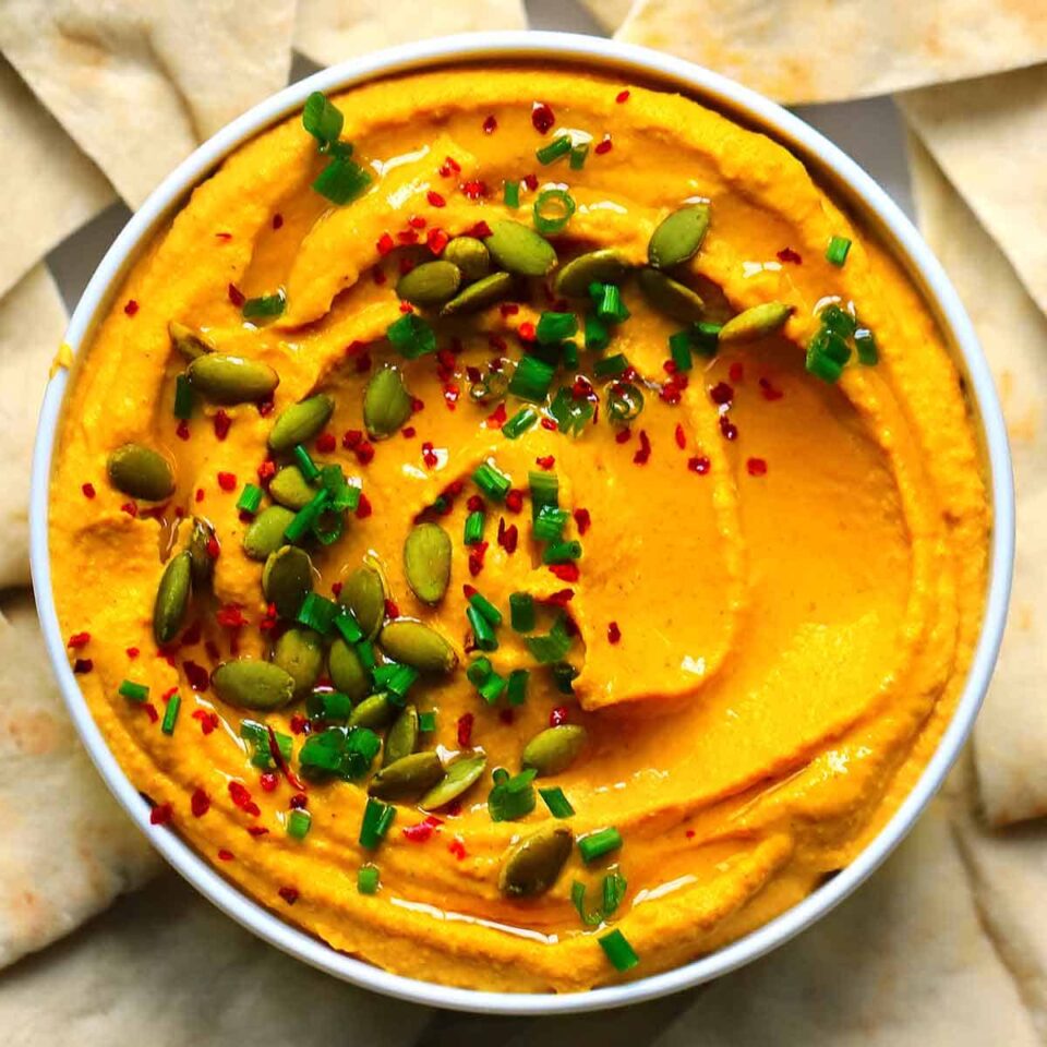 Square close up photo of an overhead view of pumpkin hummus with curry topped with chives, pumpkin seeds, olive oil, and Aleppo pepper.