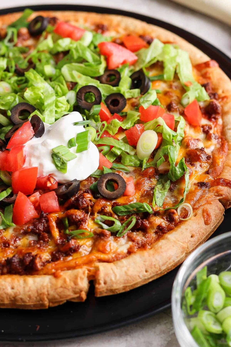 Taco pizza topped with shredded lettuce, sour cream, diced tomatoes, and olives.