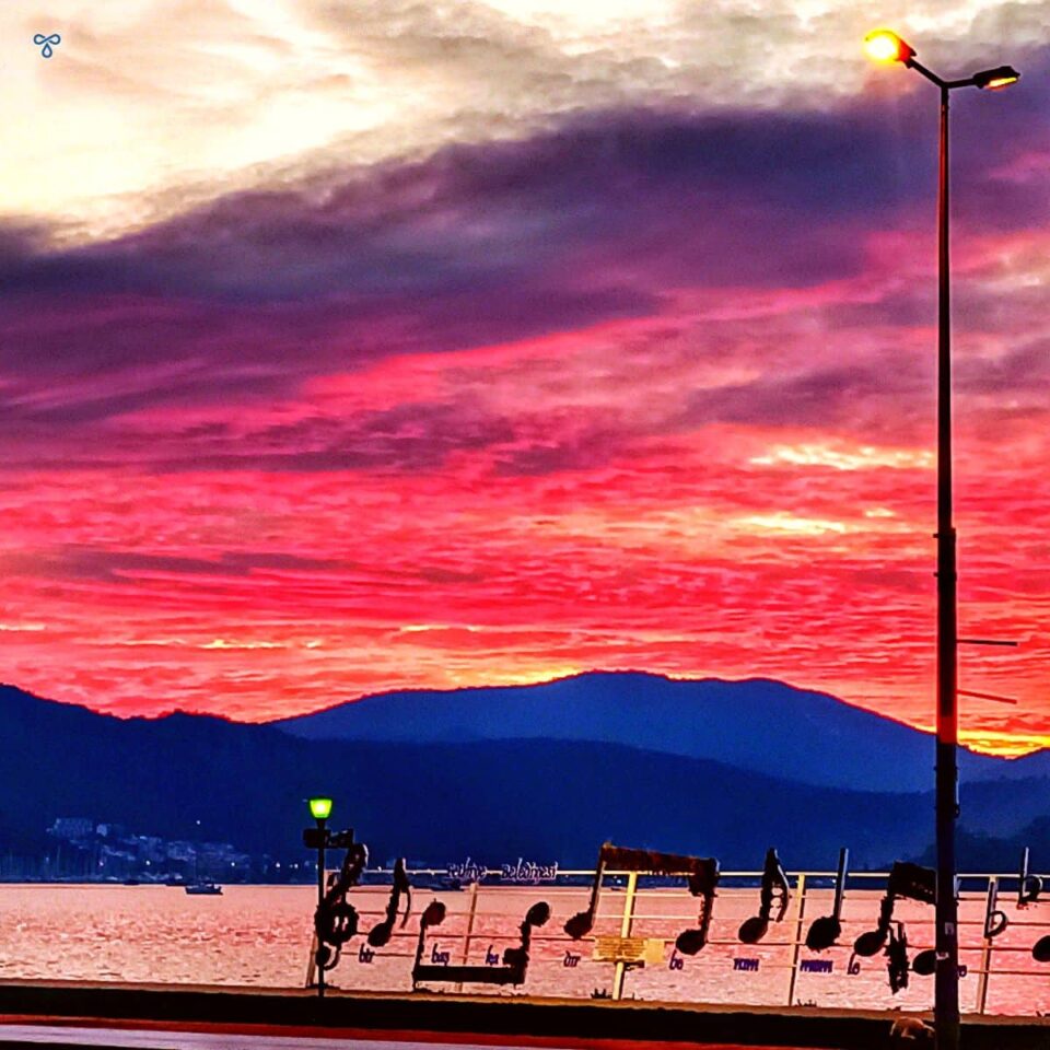 A vibrant pink cloudy sunset over hills and the bay in the foreground. A monument of a bar of a music is on the shoreline.