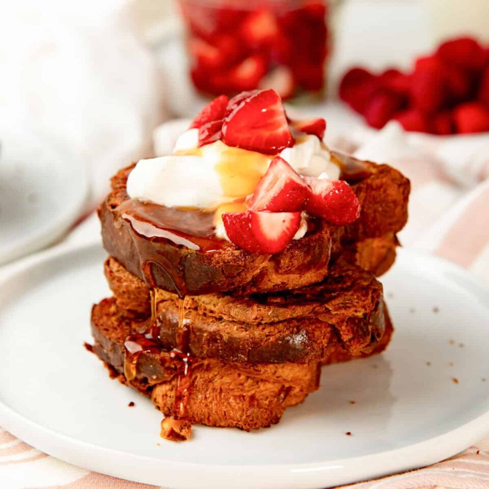 French toast slices on a white plate with whipped cream, strawberries and maple syrup on top.