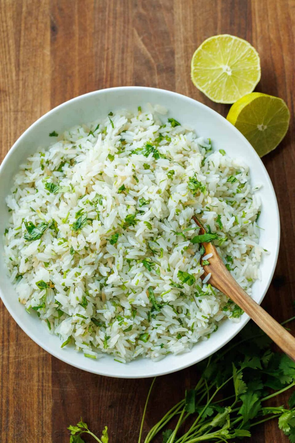 Cilantro lime rice in a large white serving bowl with a wooden spoon and slice limes