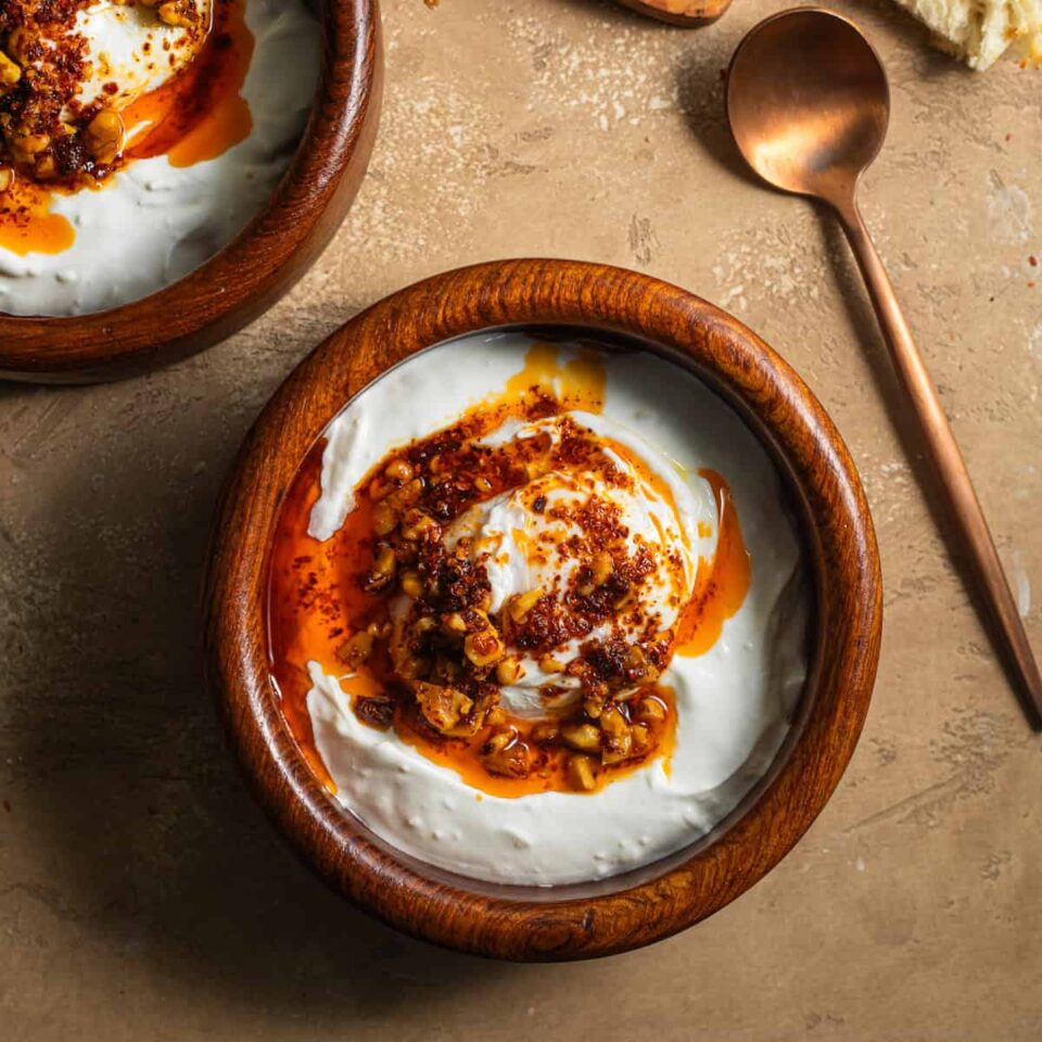 A wooden bowl with yogurt, a poached egg, and a spiced oil with walnuts and a spoon in the background.