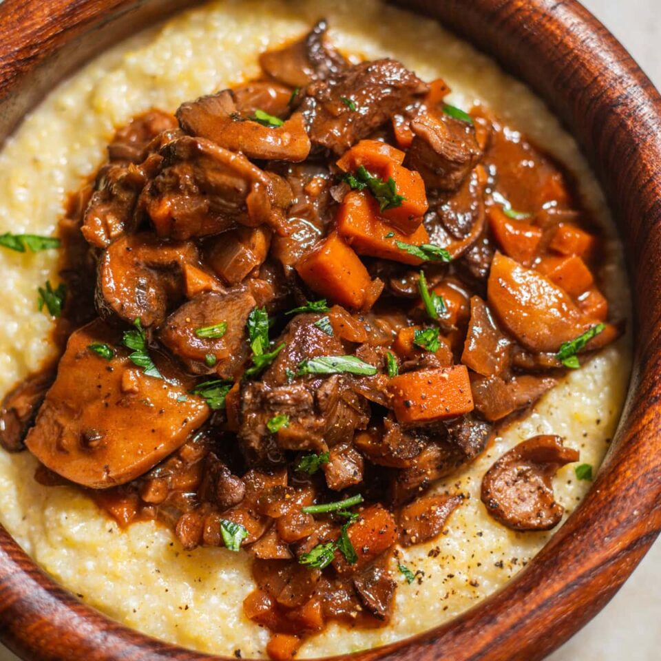 Creamy polenta topped with mushroom bourguignon and fresh parsley in a wooden bowl.