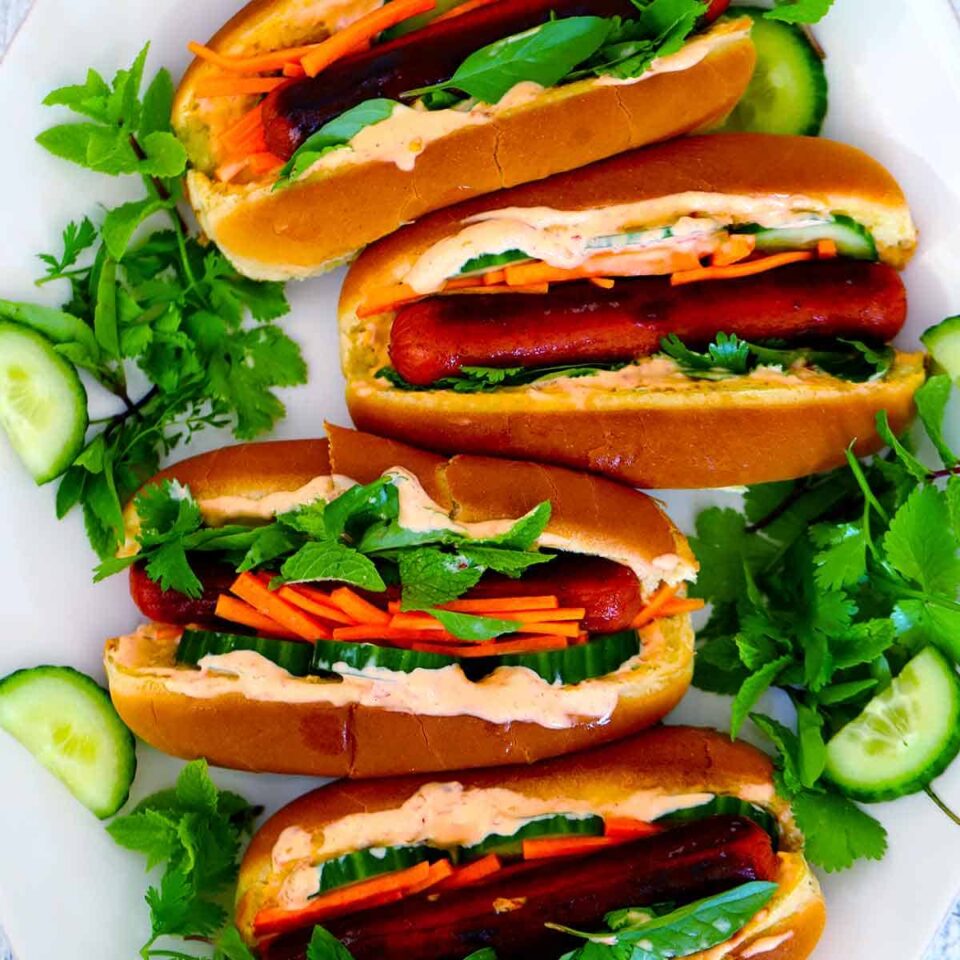 Square photo of Bánh Mì hot dogs, overhead view, surrounded by fresh mint, basil, and cilantro with cucumber slices.