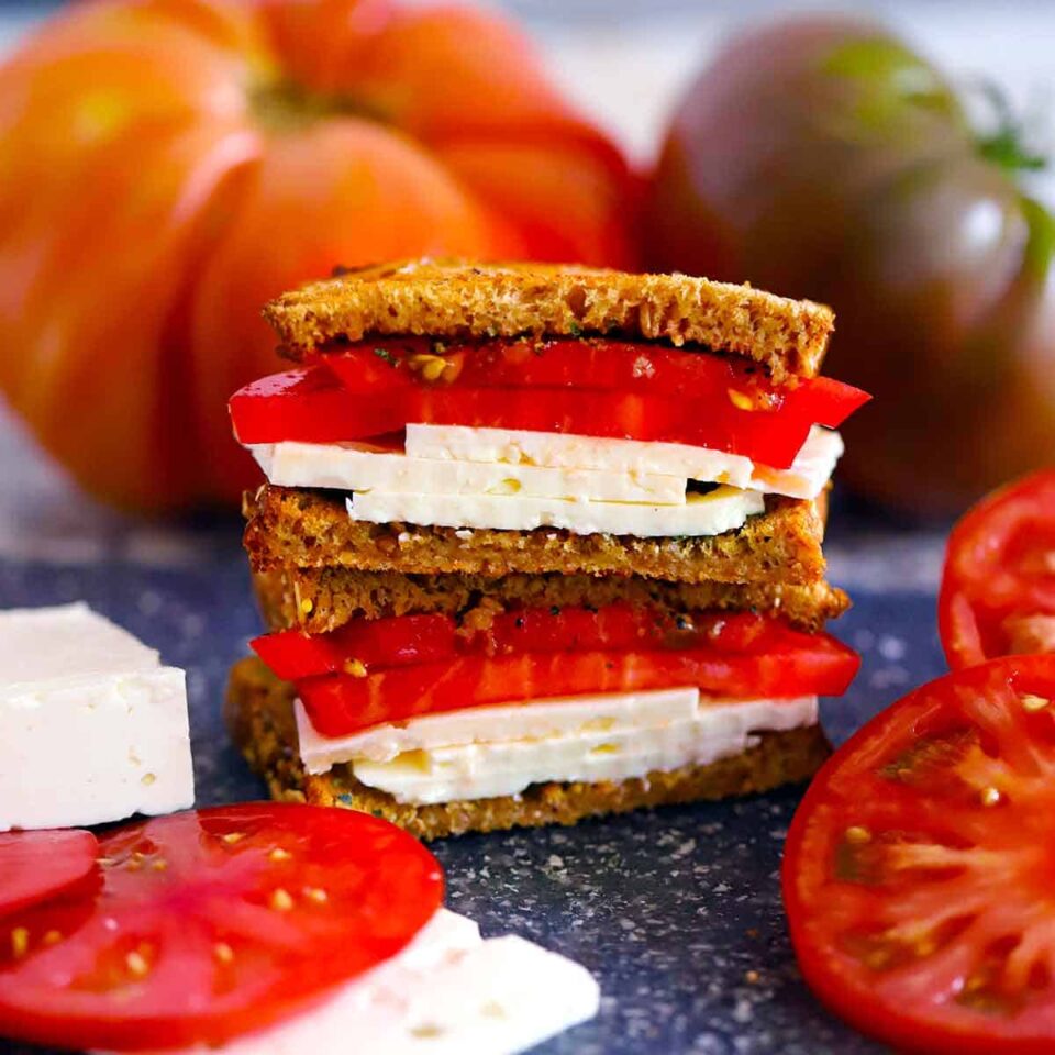 Square photo of two halves of a feta and tomato sandwich stacked on a cutting board surrounded by sliced feta and tomatoes.