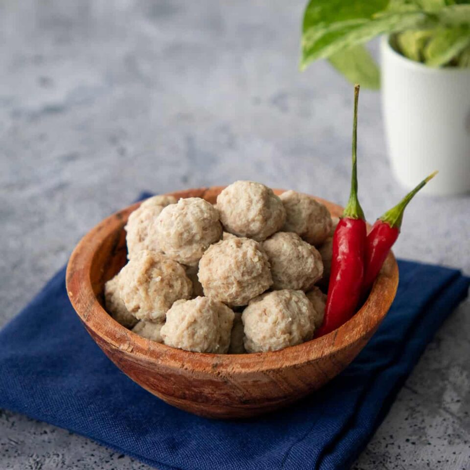 a bowl of moo deng meatballs with 2 chilies and a plant in the background