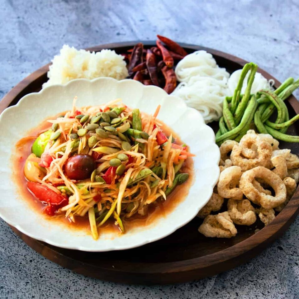 papaya salad lao style on a plate with a side of crispy pork rind, long beans, rice noodles, dried chilies and sticky rice